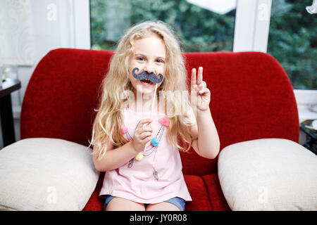 Jolie petite fille avec des moustaches de papier alors qu'il était assis sur le fauteuil rouge à la maison. Banque D'Images