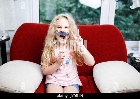 Jolie petite fille avec des moustaches de papier alors qu'il était assis sur le fauteuil rouge à la maison. Banque D'Images