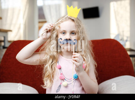 Cute little girl avec la couronne de papier et des moustaches, assis sur chaise rouge à la maison. Banque D'Images