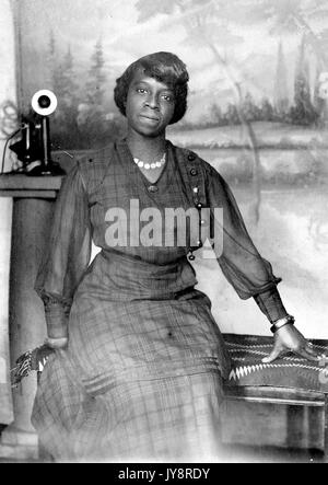 Portrait assis de trois quarts de longueur d'une femme afro-américaine mûre, assise sur un banc en lambeaux devant un fond peint et à côté d'un téléphone, portant une robe à carreaux, un collier, un bracelet, et une expression neutre, 1915.