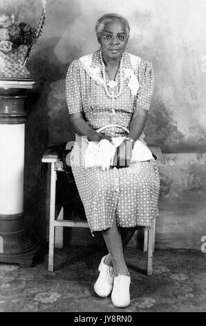 Assis sur toute la longueur portrait of elderly african american woman, assis devant un fond peint à côté d'un pilier avec un panier de fleurs, vêtue d'une robe à motifs, des verres, un collier, des boucles d'oreilles, et chaussent des chaussures, tenant un sac à main, 1915. Banque D'Images