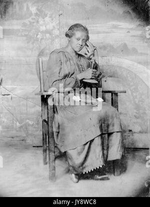 Full Length portrait of African American Woman, assis dans une chaise en bois, vêtue d'une robe sombre, au téléphone, avec une expression du visage grave, 1920. Banque D'Images