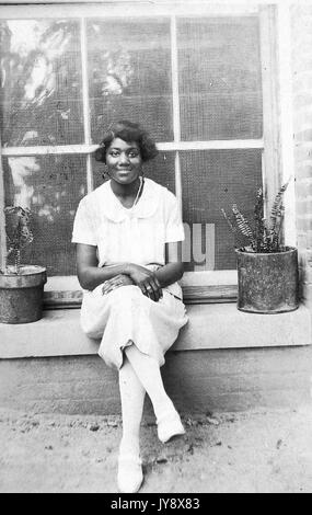 Portrait d'une jeune femme afro-américaine, à l'extérieur assis devant une fenêtre, entre deux plantes en pot, porter tout blanc, avec une expression du visage souriant, les mains sur ses genoux, 1940. Banque D'Images