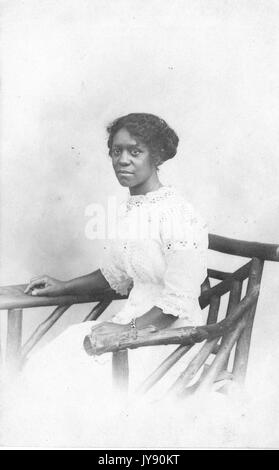 Portrait d'une femme afro-américaine assise dans une chaise en bois sculpté à la main, vêtue d'une robe blanche, à Willow Grove park, New York, 1915. Banque D'Images