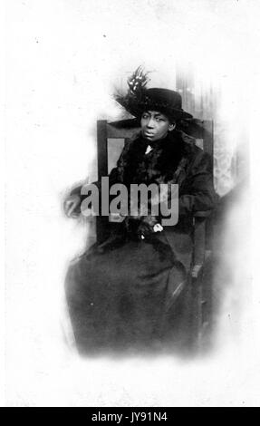 Assis pleine longueur portrait of mature african american woman avec une expression neutre, portant une robe sombre, un manteau foncé, et un chapeau à plumes, 1915. Banque D'Images