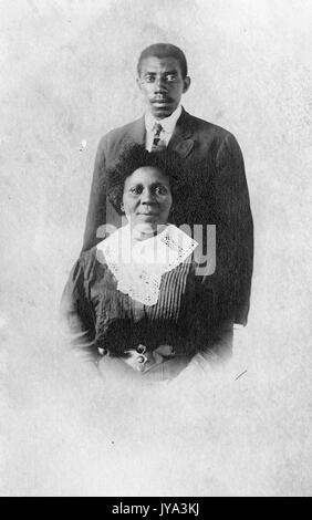 Young african american couple, un homme et une femme, la femme assise en face de l'homme portant une robe, l'homme debout et vêtu d'un costume, recadré à la photographie originale, 1932. Banque D'Images