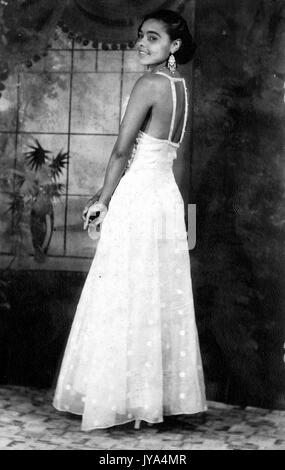 Pretty young African American girl posant pour des photographies d'une robe blanche, à la vue de derrière par dessus son épaule à la caméra et souriant, toile peinte visible, 1932. Banque D'Images