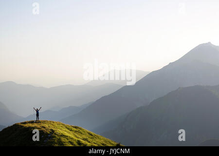 Soman, debout sur une colline couverte d'herbe avec ses mains en l'air et vue imprenable sur les montagnes au coucher du soleil, Slatnik, Slovénie. Banque D'Images