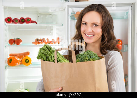 Happy Woman Holding Grocery Shopping Bag avec des légumes en face de réfrigérateur ouvert Banque D'Images