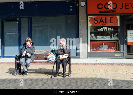 Deux vieilles femmes assises sur un banc public dans une rue piétonne pavée avec des magasins sont vides à l'arrière-plan Banque D'Images