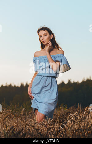 Belle jeune fille en champ de blé au coucher du soleil. Fille en robe bleue occasionnels bénéficiant d soirée ensoleillée Banque D'Images