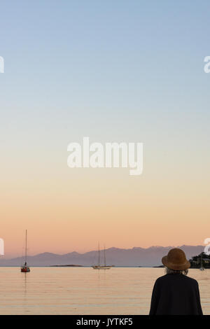 Une femme aux cheveux gris portant un caractère véritable paille vintage style helmut hat regardant les voiliers sur la baie au coucher du soleil. Banque D'Images
