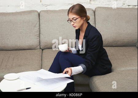 Belle jeune business woman sitting on sofa at office avec des documents et du café Banque D'Images