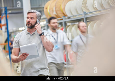 Surveillant masculin avec presse-papiers en conversation sur ell phone dans l'usine de fibre optique Banque D'Images