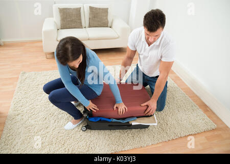 Jeune couple essayant de fermer Valise avec beaucoup de vêtements à la maison Banque D'Images