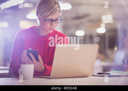 Femme sérieuse travaillant tard, using cell phone at desk in office Banque D'Images