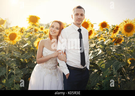 Rousse mariée mariage en robes blanches et le marié en chemise et cravate de Pentecôte dans l'étreindre permanent champ de tournesols. Avec le soleil de l'été Banque D'Images