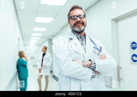 Portrait of positive doctor standing in hospital hallway, les bras croisés. Médecin mature avec des collègues parler en arrière-plan. Banque D'Images