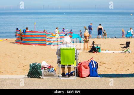 Femme âgée assise seule sur une chaise longue avec ses bagages donnant sur une plage bondée à la populaire station balnéaire de Colwyn Bay, dans le Nord du Pays de Galles sur un jour d'été pendant les vacances d'été, Pays de Galles, Royaume-Uni Banque D'Images