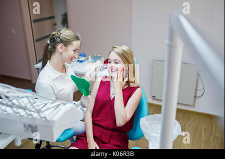 Belle dentiste montrant ses nouvelles dents du patient par le miroir dentaire en cabinet. Banque D'Images