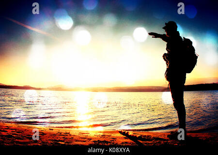 L'effet de grain de film. Homme heureux avec la main dans l'air. Grand randonneur dans les vêtements de sport sac à dos sombre avec des stands sur la plage admirer le coucher du soleil à l'horizon avec l'axe Banque D'Images