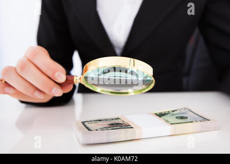 Portrait of young businesswoman scrutant dollar bills with magnifying glass at office desk Banque D'Images