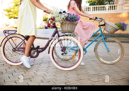 Vue latérale de l'image recadrée deux jeunes femmes en robes retro circonscription bicyclettes sur une rue de la ville Banque D'Images