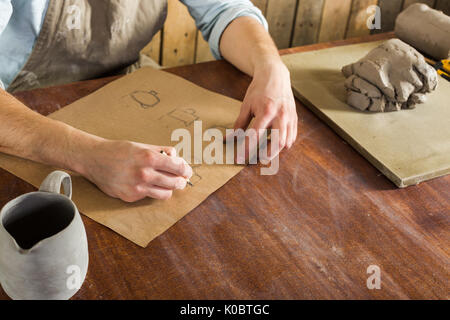 Fabrication de poterie, céramique, outils d'artisans concept art - Vue de dessus des mains du jeune homme au travail sur le dessin la faïence, un céramiste vêtu d'une chemise et d'un tablier assis derrière un bureau et pot fini. Banque D'Images