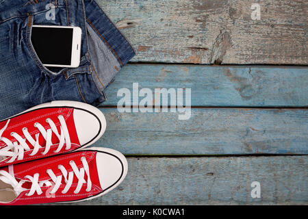 Sneakers rétro rouge et smartphone dans une poche de jeans bleu sur un fond de bois. Banque D'Images