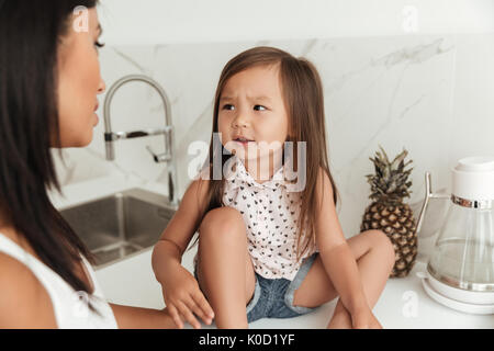 Petite fille asiatique avec les sourcils face à sa mère alors qu'à assise sur une table dans une cuisine