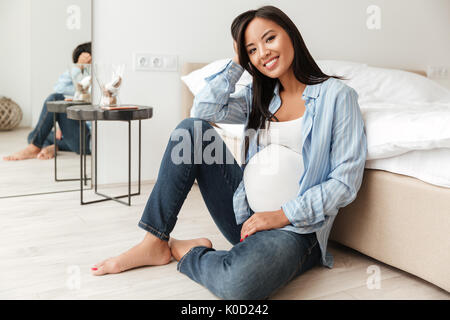 Happy pregnant asian woman leaning on un canapé tout en étant assis sur le plancher à la maison Banque D'Images