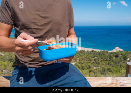 Libre d'un jeune homme de race blanche de manger une salade de lentilles à partir d'un récipient en plastique bleu à l'extérieur, avec la mer en arrière-plan Banque D'Images