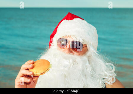 Père Noël sur la plage de manger un hamburger. Le concept d'une mauvaise alimentation. Banque D'Images