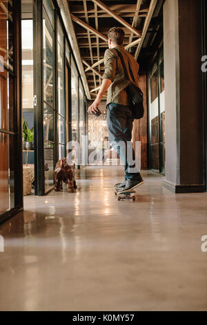 Vue arrière sur toute la longueur du coup jeune homme skateboarding in modern office. Businessman skating grâce à son bureau de démarrage. Banque D'Images