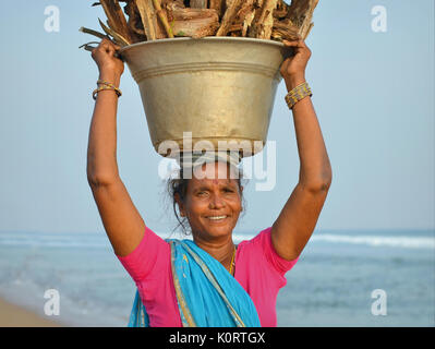 Une femme indienne âgée d'Orissan de la communauté de pêcheurs locale de Puri porte une lourde charge de bois de chauffage sur sa tête et sourit pour l'appareil photo. Banque D'Images