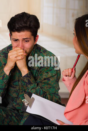 Malheureux beau jeune soldat en uniforme, d'hommes qui souffrent de stress post-traumatique après la guerre, avec les deux mains dans sa bouche, écoute le thérapeute dans un centre de santé médical, dans un contexte avec des copeaux Banque D'Images