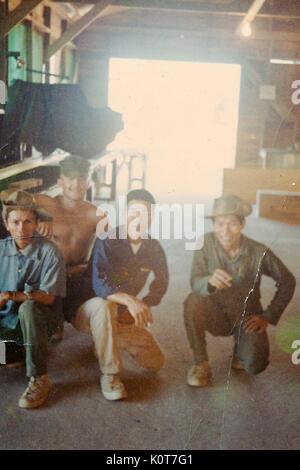 Une photographie d'un soldat de l'armée des États-Unis qui pose pour une photo avec trois hommes vietnamiens, ils sont rassemblés à l'intérieur d'un bâtiment en bois, Vietnam, 1968. Banque D'Images