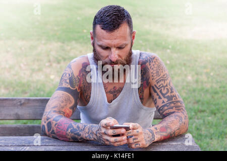 Homme barbu tatoué écrit avec smart phone dans le parc Banque D'Images