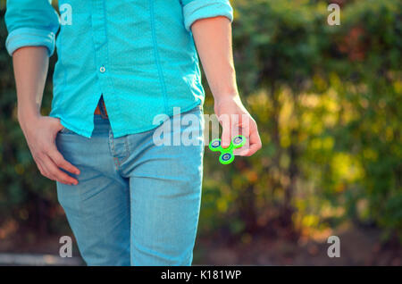 Adolescent hand holding populaires gadget antistress fidget spinner. L'homme sur l'azur shirt et jeans bleu jouant avec green spinner en plein air sur la bri Banque D'Images