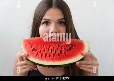 Une fille mord un morceau de melon d'eau douce. La brunette est vêtu d'un chandail noir. La jeune fille se tient sur un fond clair. Banque D'Images