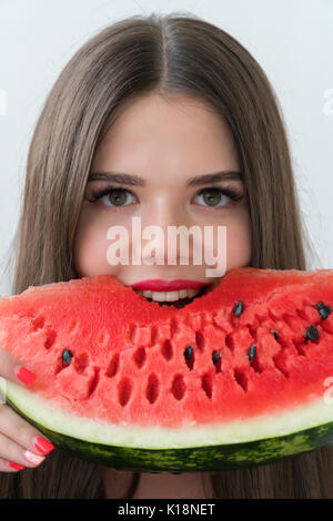 Une fille mord un morceau de melon d'eau douce. La brunette est vêtu d'un chandail noir. La jeune fille se tient sur un fond clair. Banque D'Images