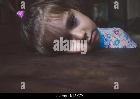 Portrait de petite fille triste. fille inclina la tête sur la table et regarde devant lui. Visage triste. L'enfant est fatigué, la tête sur une table en bois Banque D'Images