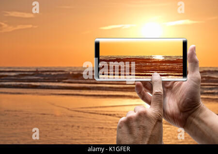 Deux mains tenant un mobile Smartphone et prendre une pictureof Coucher du soleil sur l'océan dans l'heure d'été Banque D'Images