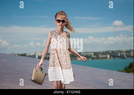 Belle petite fille en robe à la mode et des lunettes porte un sac sur fond de ciel Mer et ville Banque D'Images