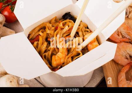 Des nouilles avec des légumes, des crevettes avec des baguettes en bois sombre sur un arrière-plan dans une boîte blanche. Banque D'Images