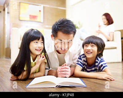 Le père et les enfants asiatiques lying on floor reading book avec la mère à l'arrière-plan.