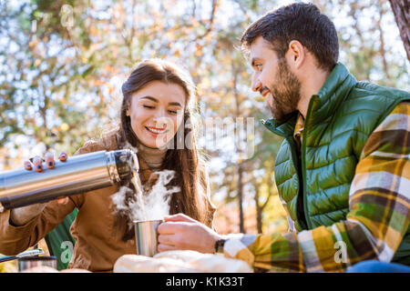 Close-up portrait of smiling young woman pouring thermos de thé chaud pour jeune homme barbu assis dehors à la journée ensoleillée d'automne Banque D'Images