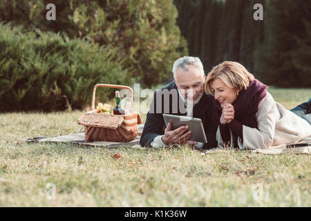 Beautiful smiling woman using digital tablet while lying on plaid avec panier à pique-nique dans le parc en automne Banque D'Images