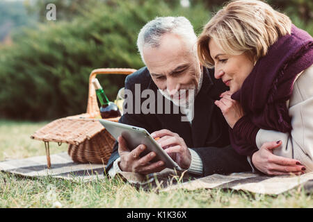 Beautiful smiling woman using digital tablet while lying on plaid avec panier à pique-nique dans le parc en automne Banque D'Images