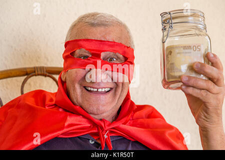 Vieil homme habillé en super héros, en souriant, assis sur chaise en osier et tenant un pot plein de billets sur fond de mur jaune Banque D'Images